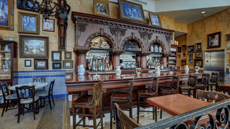 Indoor bar area with ornate dark wood counter and shelving, decorated with framed photos and ceramic pitchers, and seating with wooden chairs and small tables.