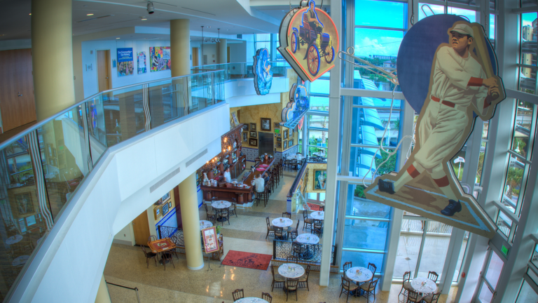 View from an upper level inside a building showing a dining area with multiple tables, a bar, large sports themed decorations, and floor to ceiling windows.