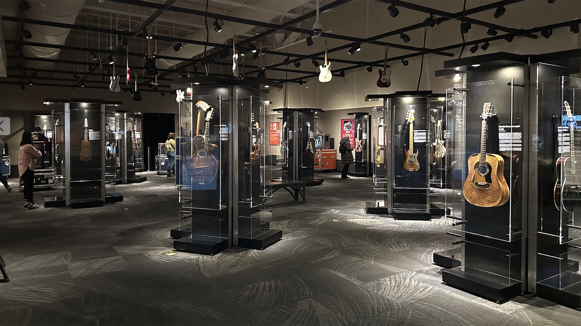 Wide view of a dimly lit guitar exhibition gallery with multiple guitars displayed in tall glass cases and hanging from the ceiling as several visitors walk and read the panels