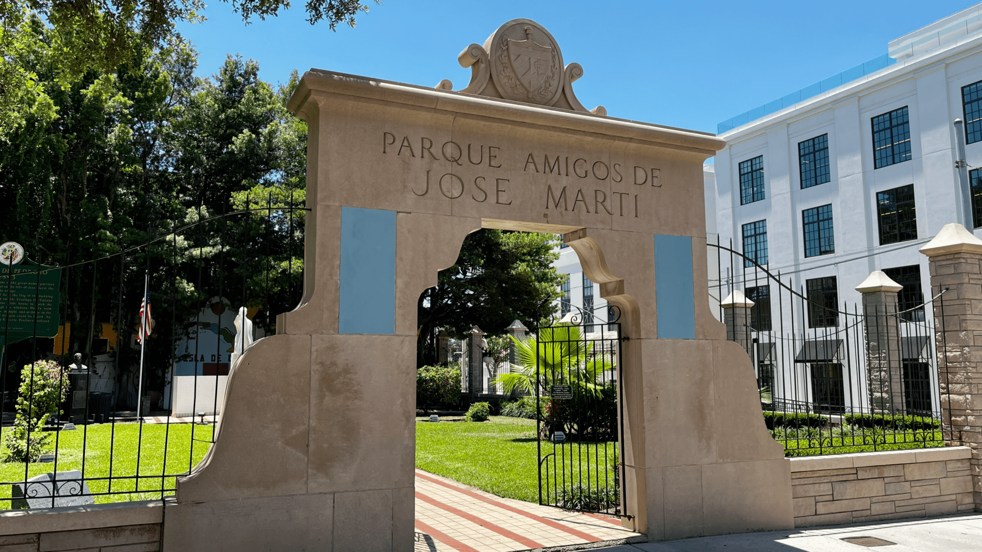 Entrance archway to Parque Amigos de José Martí in Ybor City, with the park’s name carved above an open gate, greenery inside the park and a white building in the background.